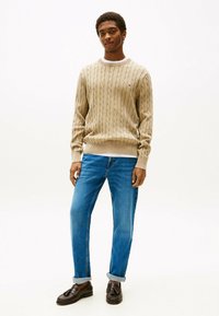 Beige kabelgebreide trui met lange mouwen, ribgebreide manchetten en kraag, gecombineerd met blauwe spijkerbroek en donkere loafers. Eenvoudig ontwerp en textuur.