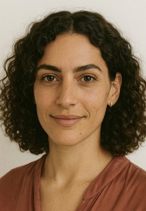 Ritratto di una persona con capelli ricci scuri e pelle chiara, che indossa una camicia rosa tenue. I tratti includono lentiggini e piccoli orecchini a cerchio.