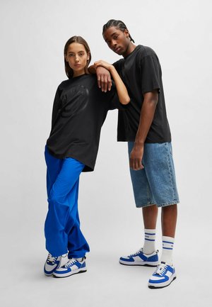 T-shirt nere oversize, pantaloni larghi blu e shorts di denim. Sneakers blu e bianche con calzini a righe, in piedi contro uno sfondo chiaro.