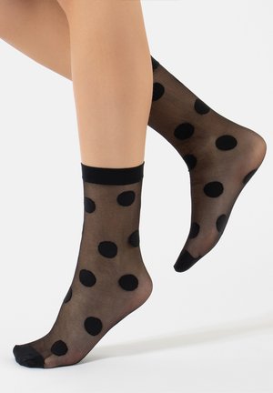 Gambe che indossano calzini neri trasparenti alla caviglia con grandi pois neri opachi su uno sfondo bianco, piedi leggermente sollevati.