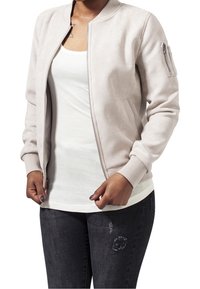 Světle béžová bomber bunda z měkké látky, s kapsou na zip na levém rukávu, ribovanými manžetami a uvolněným střihem.