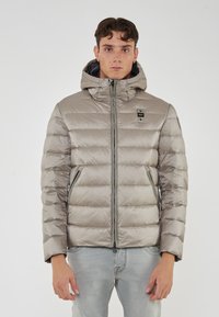 Giovane uomo che indossa un giubbotto leggero grigio con cappuccio e tasche con zip, abbinato a jeans azzurri, in piedi contro uno sfondo bianco uniforme.
