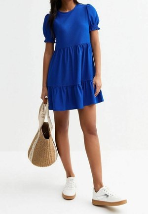 Donna che indossa un abito a balze di colore blu reale a maniche corte, sneakers bianche e tiene in mano una borsa intrecciata in paglia con manici color crema.