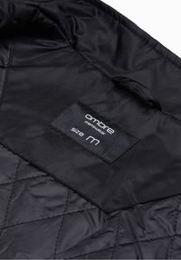 Tissu matelassé noir avec une étiquette cousue portant l'inscription "ombre menswear size M" et une petite boucle de suspension au-dessus de l'étiquette.