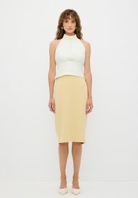 Halter krční top bílý, přiléhavý v pase, spárovaný s béžovou sukní do půli stehenní ve stylu tužky. Modelka má na sobě krémové špičaté boty.