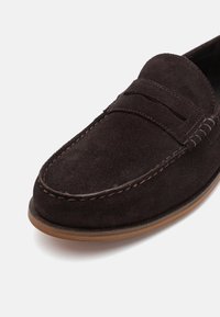 Mocassins en suède marron foncé avec une couture et un détail de bandeau "penny". Texture lisse, bout arrondi et semelle en caoutchouc marron clair.