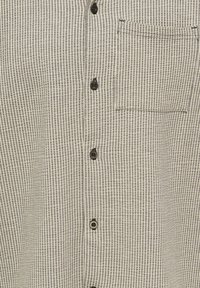 Chemise boutonnée à rayures verticales beige et grises avec des boutons noirs et une poche poitrine sur le côté droit.