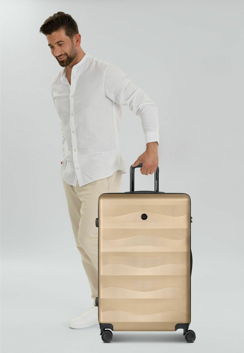 Homme en chemise blanche et pantalon beige tenant la poignée d'une grande valise rigide beige à quatre roulettes.