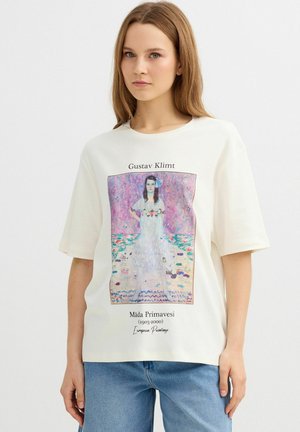 Joven mujer vistiendo una camiseta blanca con una impresión colorida de una pintura de Gustav Klimt y vaqueros azules sobre un fondo claro y sencillo.