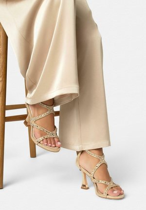 Femme portant un pantalon beige à jambes larges et des sandales dorées à talons hauts avec des ornements scintillants, assise sur une chaise en bois.