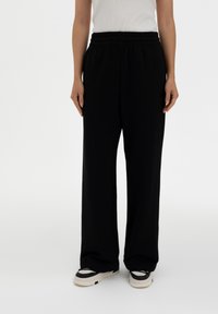 Black wide-leg sweatpants with an elastic waistband and subtle logo detail, made from soft fabric. Styled with casual sneakers.