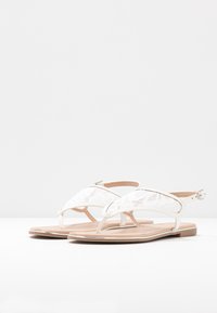 Valkoiset sandaalit, joissa on pitsikoristeet, T-nauhan muotoilu ja säädettävä nilkk buckle. Tasapohja, jossa on vaaleanbeige sisäpohja.