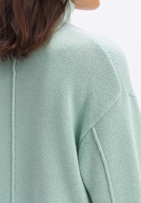 Vaaleanvihreä neulepaita, jossa on korkea kaula-aukko, roikkuvat olkapäät ja teksturoitu kangas, jossa on keskisauma selässä.