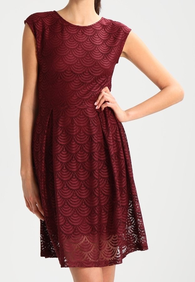 Robe sans manches en dentelle bordeaux avec un motif festonné, taille cintrée et jupe évasée longueur genou portée par une personne avec une main sur la hanche.
