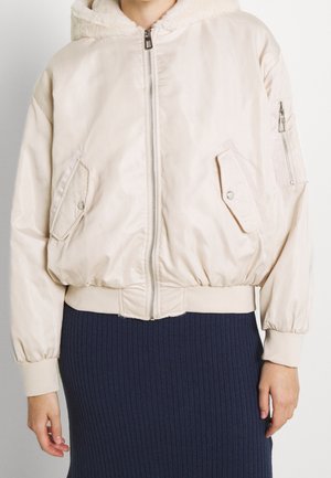Beżowa kurtka typu bomber z kapturem wykończonym sztucznym futrem, zamknięciem na zamek, trzema kieszeniami oraz elastycznymi ściągaczami. Gładka tkanina o lekko błyszczącym wykończeniu.