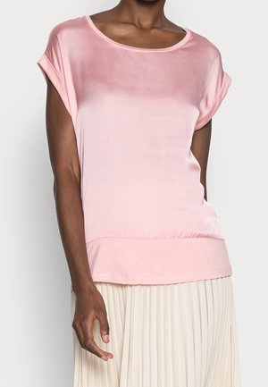 Femme vêtue d'un chemisier en satin rose à manches courtes avec des poignets retroussés et d'une jupe plissée blanche, vue de l'épaule aux hanches.