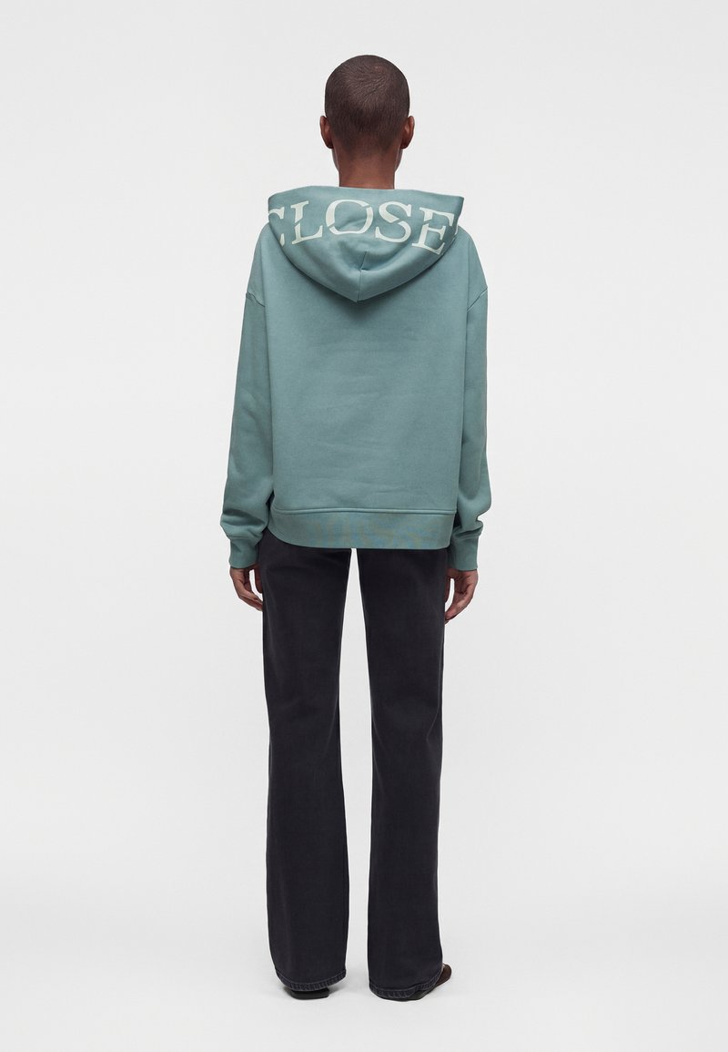 Personne debout de dos, portant un sweat à capuche vert avec "CLOSE" sur la capuche et un pantalon noir, sur un fond blanc uni.