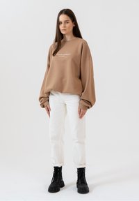 Brązowa, oversize bluza z białym tekstem, zestawiona z białymi dżinsami i czarnymi sznurowanymi botkami. Gładki materiał, luźny krój, opadające ramiona.
