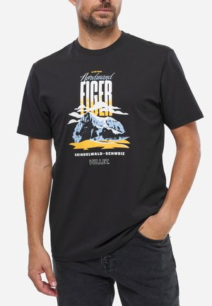 Homme portant un t-shirt noir avec un graphique de la montagne Eiger, des nuages et le soleil ; le texte indique "Nordwand Eiger 3970m Grindelwald Schweiz Millet."