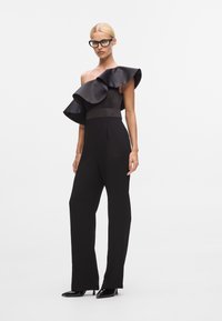 Svart jumpsuit med enaxel-design, med en stor volangdetalj. Slät tyg, vida ben och en figursydd midja med en matt finish.