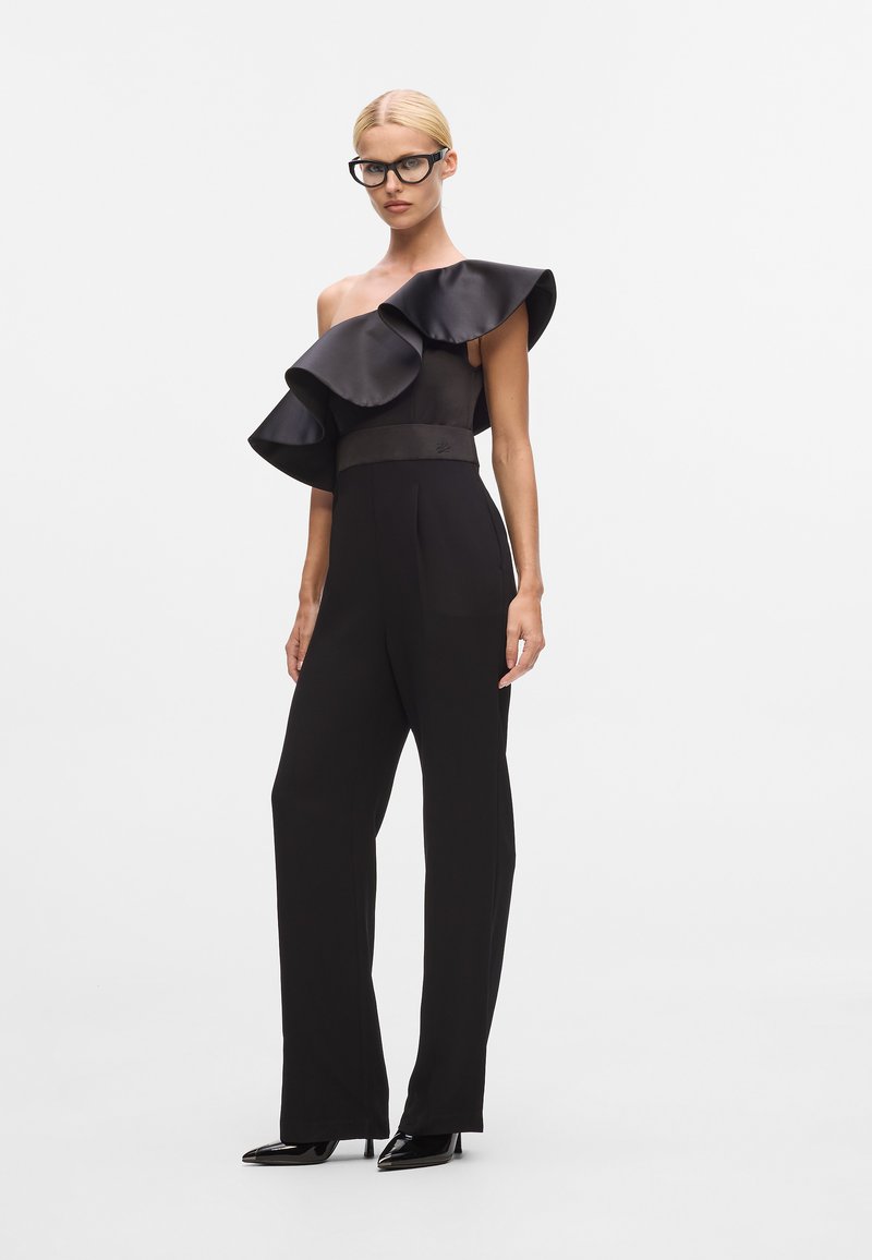 Svart jumpsuit med enaxel-design, med en stor volangdetalj. Slät tyg, vida ben och en figursydd midja med en matt finish.