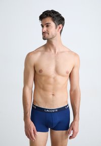 Marineblauwe boxershorts van zachte stof, met een contrasterende zwarte tailleband waarop het "LACOSTE" logo prominent is weergegeven.