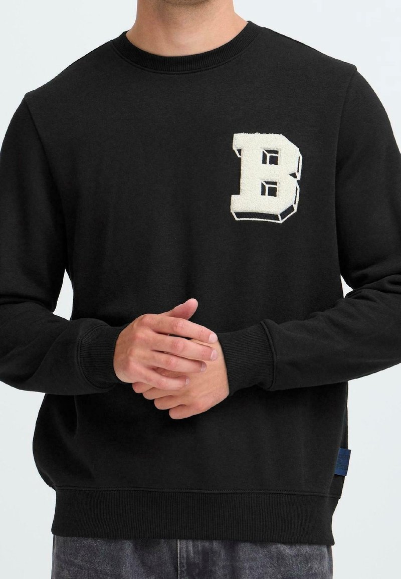Homme portant un sweat-shirt noir à col ras du cou avec la lettre "B" blanche en relief 3D sur le côté gauche de la poitrine, les mains jointes devant.