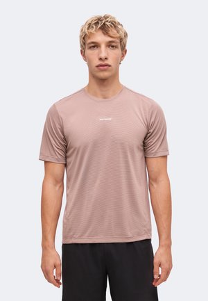 Jeune homme aux cheveux blonds courts portant un t-shirt Salomon marron clair et un short noir, debout devant un fond clair uni.