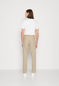 Chemise blanche à manches courtes associée à un pantalon beige à jambe droite, en tissu texturé, et des baskets blanches, vue de dos sur fond uni.