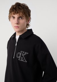 Jaunas vyras su šviesiai rudais plaukais, dėvintis juodą ketvirtį užtrauktuko megztinį su baltu "cK" logotipu ant paprasto šviesaus fono.