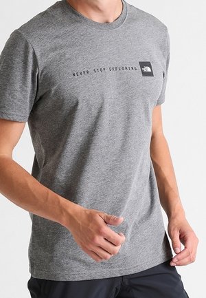 Mężczyzna noszący szarą koszulkę The North Face z napisem „Never Stop Exploring” na piersi, stojący na jednolitym jasnym tle.