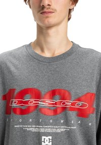 Un jeune homme portant un t-shirt gris avec un grand "1994" rouge et le texte "DCSCO SPORTSWEAR" en blanc imprimé sur le devant.