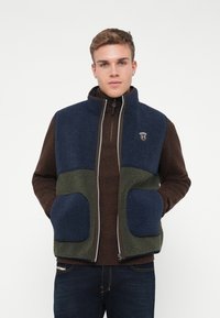 Fleece vesta s námorníckou modrou hornou časťou, tmavo hnedou spodnou časťou a zelenými akcentmi. Obsahuje zips a dve predné vrecká.