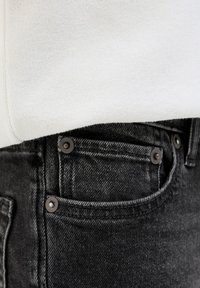 Primer plano del bolsillo de vaqueros negros con remaches metálicos, parcialmente cubierto por una tela blanca texturizada.