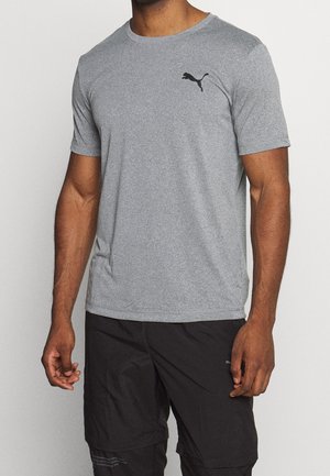 Mand iført en enkel grå Puma T-shirt og sorte atletikshorts står mod en ensfarvet baggrund.