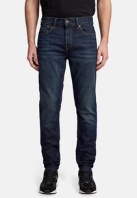 Blå denimjeans med slim fit, med fem fickor, en metallknappsgylf och kontrasterande sömmar. Bärs med svarta sportskor.