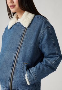 Chaqueta de denim con un suave cuello de borrego en color crema. Cuenta con un cierre frontal, bolsillos laterales y un corte relajado. Textura de denim azul.