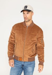 Bruine corduroy bomberjack met ribgebreide manchetten en zoom, voorrits, zijzakken en een logo label op de onderste zoom. Draag het met een beigekleurige pet.