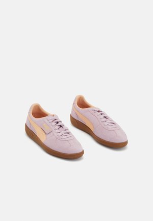 Liliowe sneakersy z zamszu z brzoskwiniowymi paskami z logo Puma i brązowymi gumowymi podeszwami, ustawione ukośnie obok siebie na białym tle.