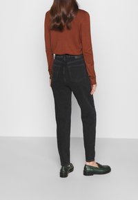 Žena stojící zády k fotoaparátu, oblečená v rustikálním dlouhém rukávu, černých džínách s vysokým pasem a tmavozelených kožených mokasínech na jednobarevném pozadí.