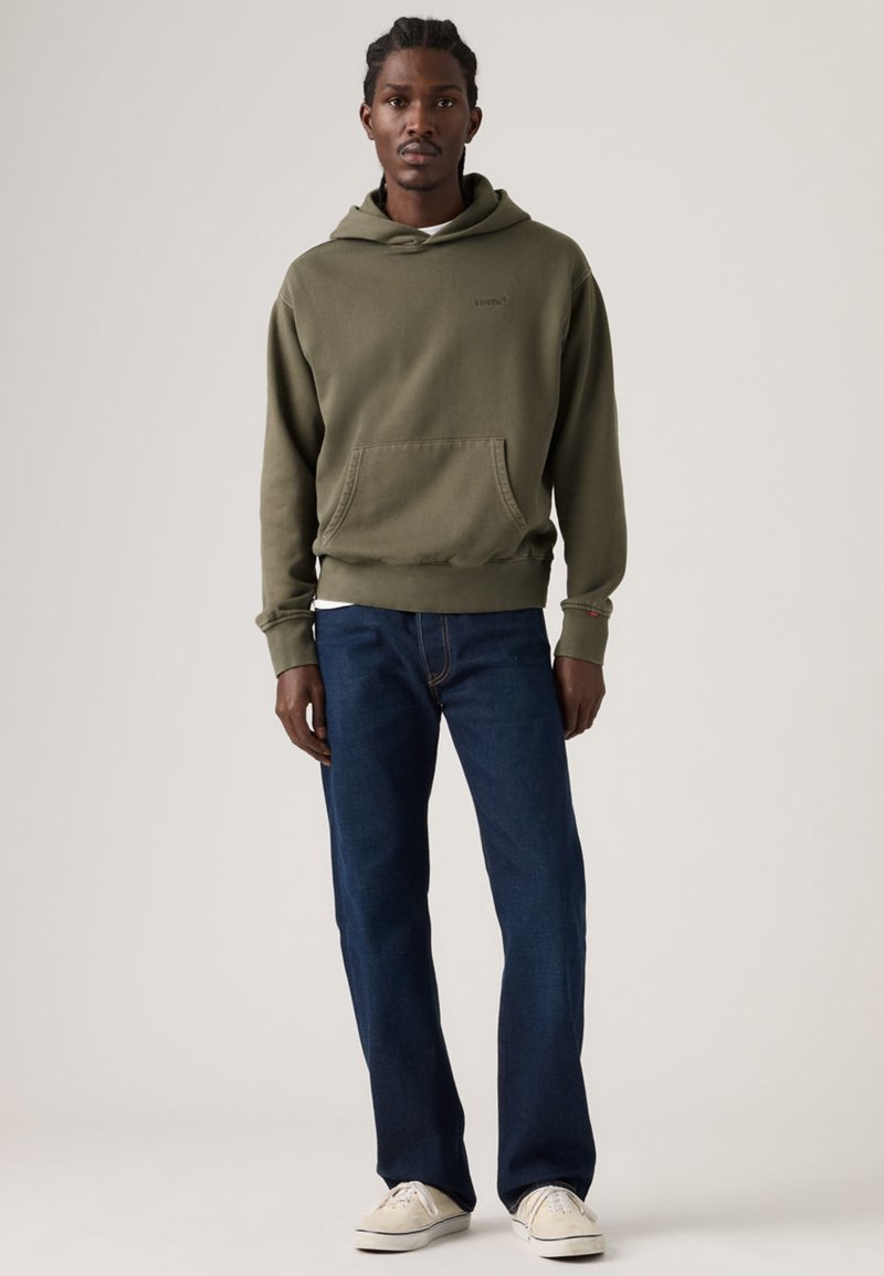 Olive green hoodie with front pocket, fitted cuffs, and logo on chest, paired with dark blue straight-leg jeans and beige sneakers.