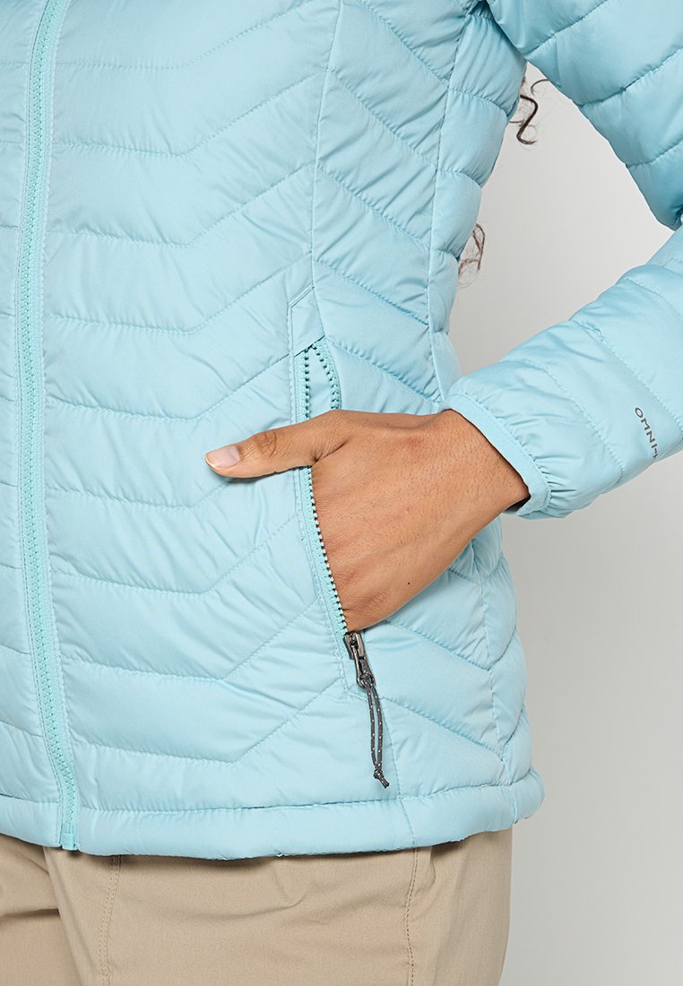 Chaqueta acolchada de color azul claro con un bolsillo con cremallera, puños acanalados y un acabado suave. El patrón de flechas en la tela añade textura.