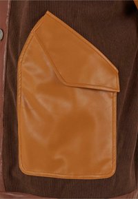 Braunes Cordstoffgewebe mit einer großen, rechteckigen Tasche aus beige Leder, die einen abgerundeten Klappenverschluss hat. Oberflächenstrukturen sind sichtbar.