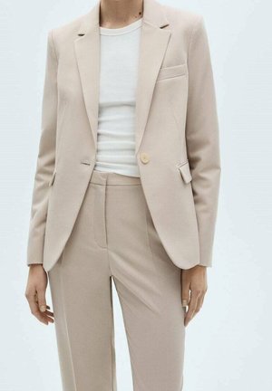 Person iført en beige skræddersyet blazer og matchende bukser over en hvid ribstrikket rundhalsede trøje, stående mod en ensfarvet baggrund.