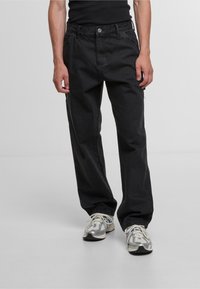 Zwarte denim broek met een relaxed fit, voorzien van een knoopsluiting en zijzakken. Draagt bij grijze sneakers met witte accenten.