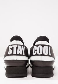 Svarta och vita sneakers med en texturerad ovandel, en kraftig klack och orden "STAY COOL" tryckta framträdande på baksidan.