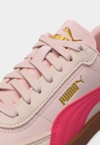 Κοντινό πλάνο ροζ αθλητικού παπουτσιού Puma με χρυσό λογότυπο, ροζ κορδόνια, υφές από σουέτ και δέρμα, και έντονη ροζ ρίγα σε λεπτομέρεια.