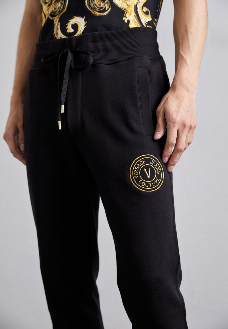 Pantalones deportivos negros con cintura elástica, cordón ajustable, bolsillos laterales y logotipo de Versace Jeans Couture bordado en dorado.