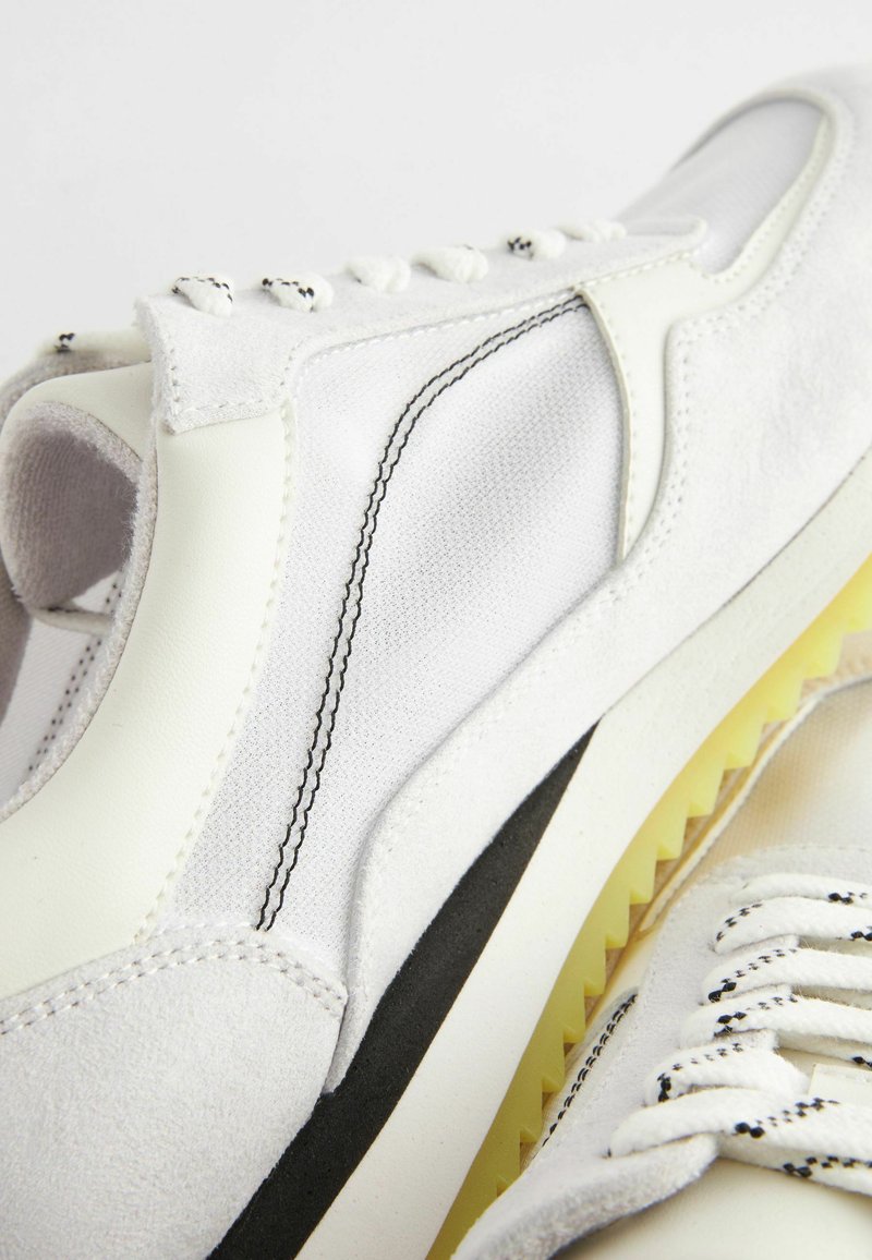 Weiße Sneaker mit einer Mischung aus glatten und strukturierten Stoffen, schwarzen Akzenten, einer leichten Sohle und kontrastierenden Nähdetails.
