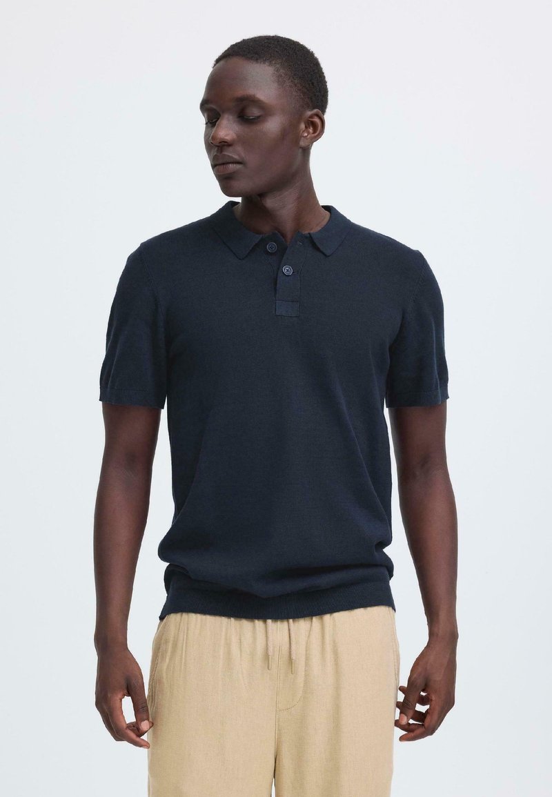 Polo bleu marine à manches courtes en tissu tricoté avec col et patte de boutonnage à deux boutons, associé à un pantalon beige à cordon de serrage.